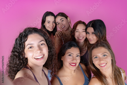 A close-knit group of women taking a joyful selfie together, set against a pink backdrop. 