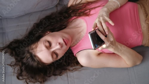 Young woman in braces with long curly hair lies on the sofa, scrolls social networks on her phone in a pink top and falls asleep, top view photo