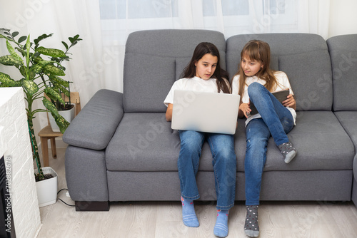 Cute little girl and older sister playing together smiling and having bonding time using a laptop on couch at home. Happy family Siblings relationship and digital technology lifestyle concept.