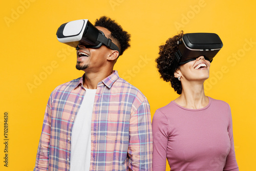 Young fun couple two friends family man woman of African American ethnicity wear purple casual clothes together watching in vr headset pc gadget look aside isolated on plain yellow orange background.