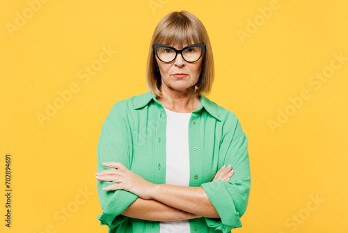 Elderly sad frowning blonde woman 50s years old wears green shirt glasses casual clothes hold hands crossed folded look camera isolated on plain yellow background studio portrait. Lifestyle concept. photo