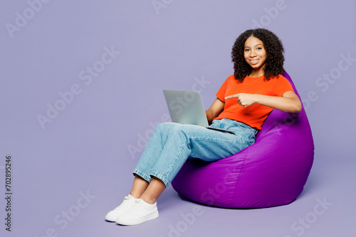 Full body kid teen girl of African American ethnicity wear orange t-shirt sit in bag chair work hold use point on laptop pc computer isolated on plain purple background. Childhood lifestyle concept.