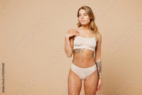 Young nice lady woman with slim body perfect skin wears nude top bra lingerie stand put hand on neck breast looking aside isolated on plain pastel light beige background. Lifestyle diet fit concept.
