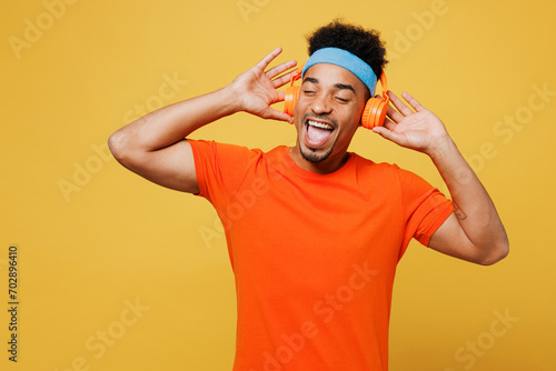 Young smiling fitness trainer instructor sporty man sportsman wearing orange t-shirt headphones listen music spend time in home gym isolated on plain yellow background. Workout sport fit abs concept.