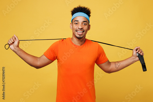 Young happy fitness trainer instructor sporty man sportsman wears orange t-shirt hold skipping rope on neck spend time in home gym isolated on plain yellow background. Workout sport fit abs concept.