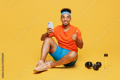 Full body young fitness trainer sporty man sportsman wear orange t-shirt sit hold mobile cell phone show thumb up train in home gym isolated on plain yellow background. Workout sport fit abs concept.