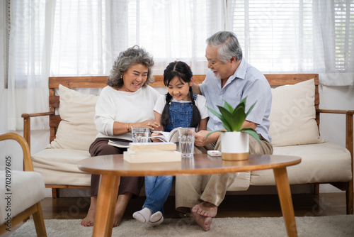 Asian senior couple watching granddaughter reading a book on sofa. Little girl visit her grandparents on holidays weekend. Love and bonding. Healthy family insurance photo
