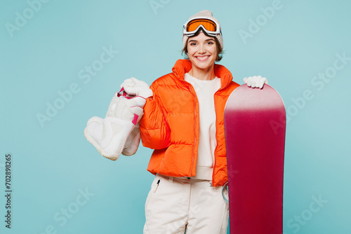 Young happy woman wear warm padded windbreaker jacket hat ski goggles mask hold snowboard ice skates look camera travel rest spend weekend winter season in mountains isolated on plain blue background.