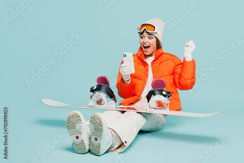 Full body winner young woman wear warm windbreaker jacket ski goggles mask sit with snowboard, mobile cell phone travel rest spend weekend winter season in mountains isolated on plain blue background.