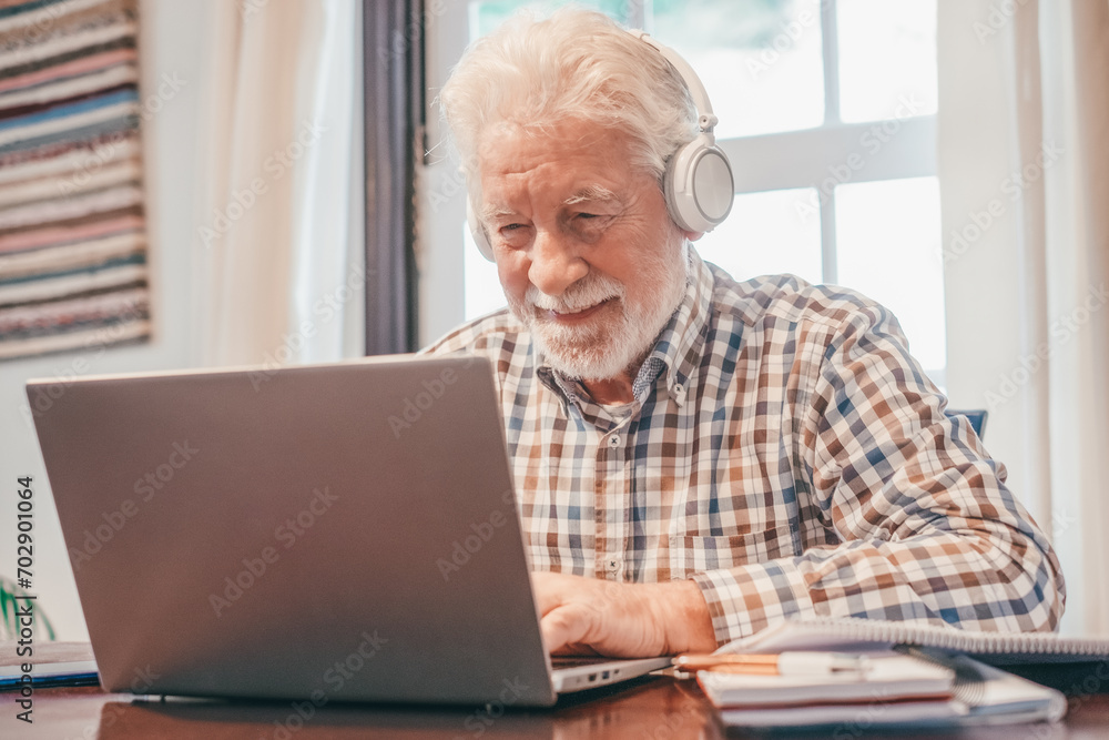 Smiling senior bearded man wearing headphones sitting at table with laptop following online course. Elderly man in checkered shirt enjoying teaching activity and learning using new technology Smiling senior bearded man wearing headphones sitting at table with laptop following online course. Elderly man in checkered shirt enjoying teaching activity and learning using new technology