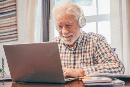 Smiling senior bearded man wearing headphones sitting at table with laptop following online course. Elderly man in checkered shirt enjoying teaching activity and learning using new technology