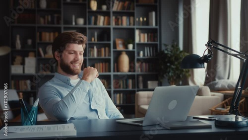 young man with beard sits at table working at laptop in home office. completes work successfully and gets great news. snap his fingers, being self contented and confident, leans back smiling