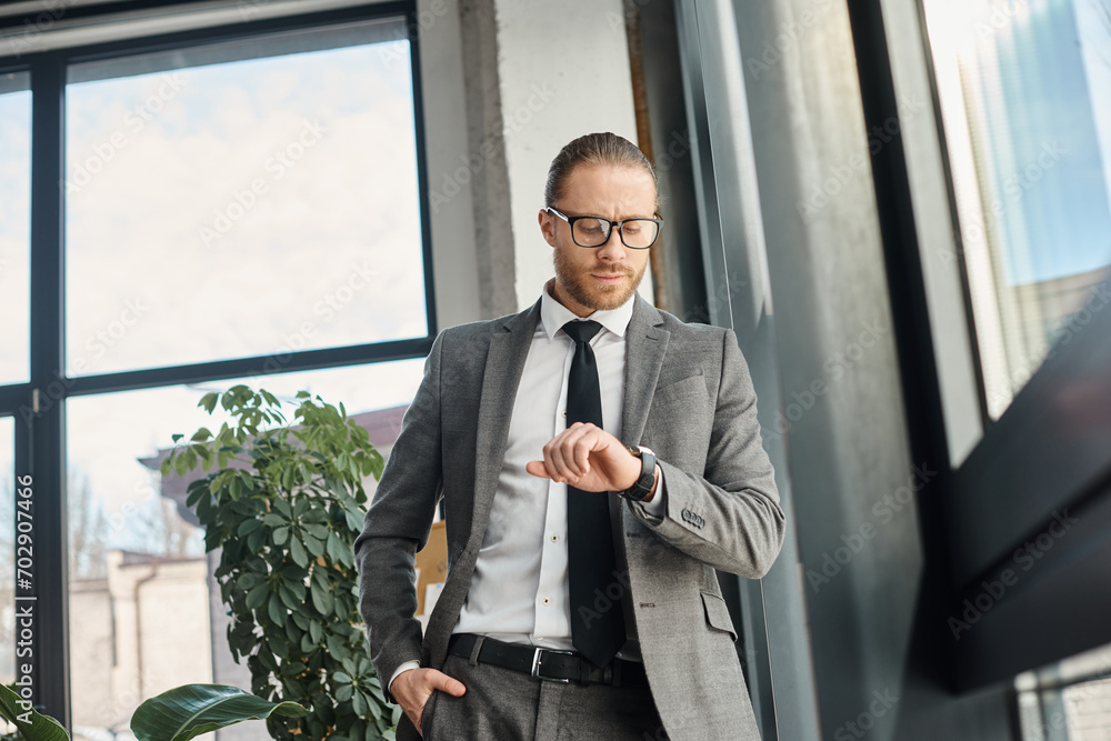 stylish businessman in grey suit and eyeglasses standing in modern office and looking at wristwatch stylish businessman in grey suit and eyeglasses standing in modern office and looking at wristwatch