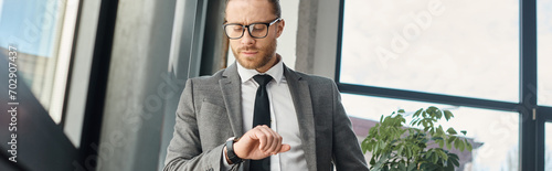 serious entrepreneur in suit and eyeglasses looking at wristwatch in office, horizontal banner