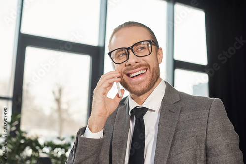 happy businessman in eyeglasses smiling during conversation on smartphone in office, portrait