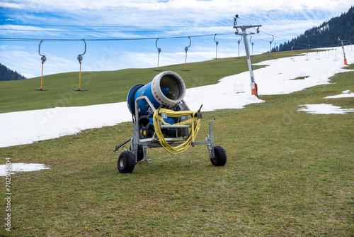 A snow cannon waiting to be used to prepare the ski trails in the ski area in Walchsee, Tyrol, Austria. A ski piste and a ski lift (T-bar lift) in the background.