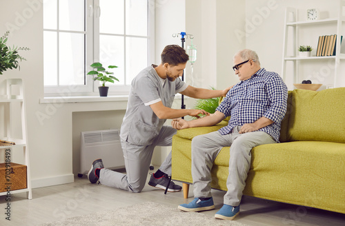 Male caregiver setting up iv drip to senior patient. Senior man sitting on sofa receiving intravenous treatment or vitamin therapy at home. Elderly healthcare, medical care, support