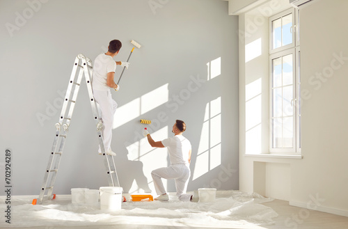 Fototapeta Naklejka Na Ścianę i Meble -  Two male workers from the professional home renovation service painting walls light gray in a big bright living room interior inside a new modern house or apartment