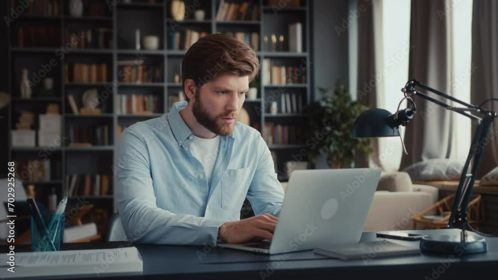 young man with beard and in blue shirt sits at table in home office and works at laptop, looks attentively with tension at screen, reads bad news, closes laptop, stands up and goes away quickly young man with beard and in blue shirt sits at table in home office and works at laptop, looks attentively with tension at screen, reads bad news, closes laptop, stands up and goes away quickly