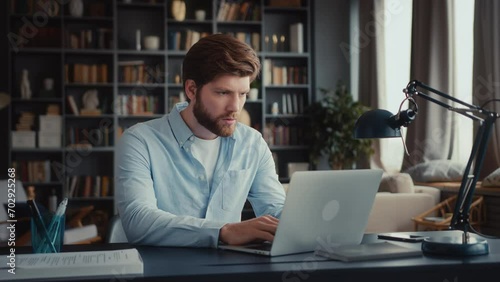 young man with beard and in blue shirt sits at table in home office and works at laptop, looks attentively with tension at screen, reads bad news, closes laptop, stands up and goes away quickly photo