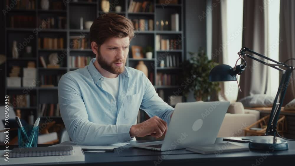 young serious and focused man sits at table in home office and works at laptop, looks at screen attentively, gets disappointed news, closes laptop frustrated, shaking head. young serious and focused man sits at table in home office and works at laptop, looks at screen attentively, gets disappointed news, closes laptop frustrated, shaking head.