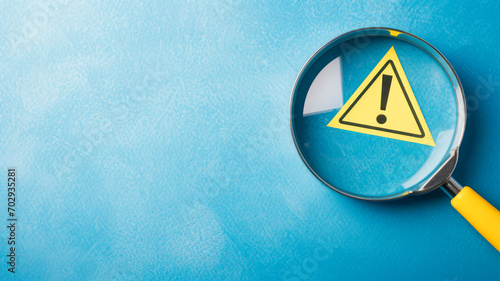 A yellow warning triangle inside the glass of a magnifying glass against a blue background, with copy space, signifies a maintenance alert and emphasizes the risk concept. photo