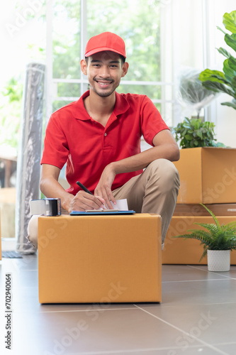 Portrait of young Asian man house moving service worker in uniform doing home relocation, checking list cardboard boxes for preparing to move. Smiling and looking at camera © pitipat