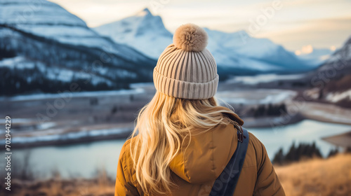 Young woman with blonde hair standing on an Icelandic dry grass field in winter, looking at the snowy mountains and lake or river landscape, wearing a yellow jacket, outdoors winter season, rearview