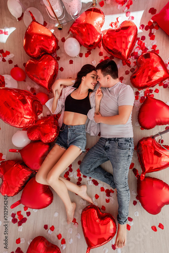 Top view of cheerful young couple lying on floor near red white balloon heart shaped, rose petals scattered around. Valentines Day romantic decorations. Man and woman looking at each other, smiling