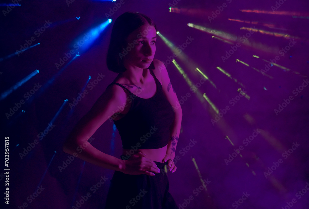 A young woman in black casual clothes poses in a studio with smoke, purple light and beams of multicolored light. The dancer demonstrates elements of experimental hip hop style dance choreography. A young woman in black casual clothes poses in a studio with smoke, purple light and beams of multicolored light. The dancer demonstrates elements of experimental hip hop style dance choreography.