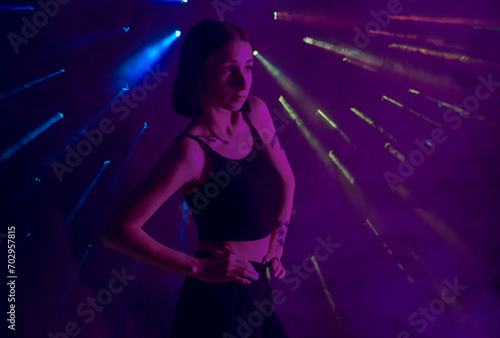 A young woman in black casual clothes poses in a studio with smoke, purple light and beams of multicolored light. The dancer demonstrates elements of experimental hip hop style dance choreography. photo