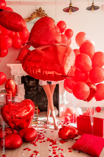 Faceless young woman standing in room filled with mix of red balloons, with romantic white fireplace. Large foil heart covered face, only long beautiful legs were visible. Valentines day, blogger