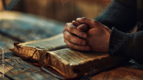 Person s hands folded in prayer over an open  well-worn bible  resting on a wooden table