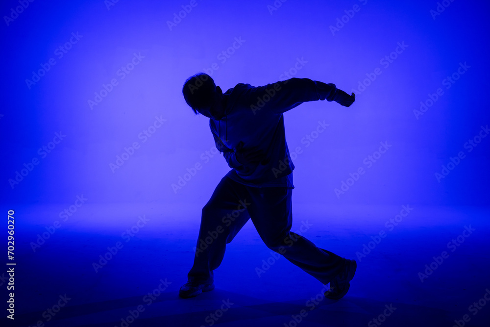 Silhouette of guy in casual clothes dancing elements of hip hop in studio with blue light. Dancer demonstrates body plasticity. Full height. Modern street choreography. Silhouette of guy in casual clothes dancing elements of hip hop in studio with blue light. Dancer demonstrates body plasticity. Full height. Modern street choreography.