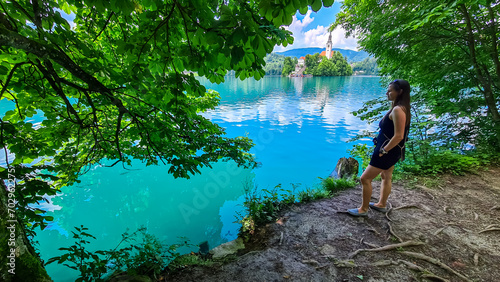 Tourist woman with panoramic view of St Mary Church build on small island on alpine lake Bled, Upper Carniola, Slovenia. Serene landscape in Julian Alps in winter. Hills covered with lush green forest