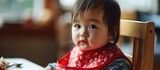 Asian toddler wearing red bib patiently seated in baby chair, awaiting meal.