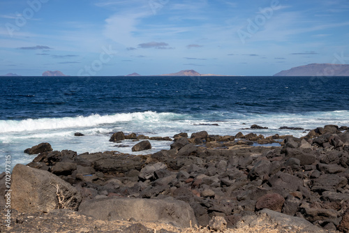 Detail of a textured dark brown to black volcanic stones on the coast of Atlantic ocean. Mountain in the background. Blue sky with some white clouds. Central Lanzarote, Canary Islands, Spain.