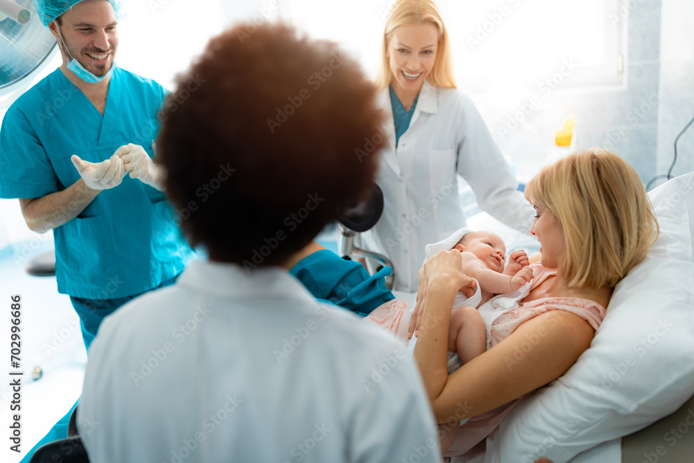 Obraz premium Beautiful young mother holding baby after giving birth in hospital surrounded by team of smiling gynecologists. Happy satisfied medical team with young woman after giving birth.
