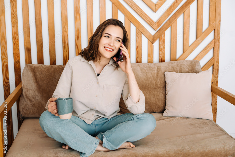 Cheerful Young Woman Talking on Phone, Relaxing on Sofa, Enjoying a Refreshing Drink in Leisure Time. Cheerful young female calls by phone, speaks, sits on sofa, enjoys cup of drink in free time. Cheerful Young Woman Talking on Phone, Relaxing on Sofa, Enjoying a Refreshing Drink in Leisure Time. Cheerful young female calls by phone, speaks, sits on sofa, enjoys cup of drink in free time.
