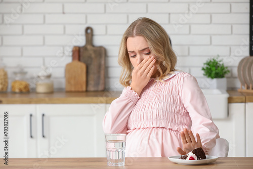 Young pregnant woman with piece of cake suffering from toxicosis at table in kitchen photo