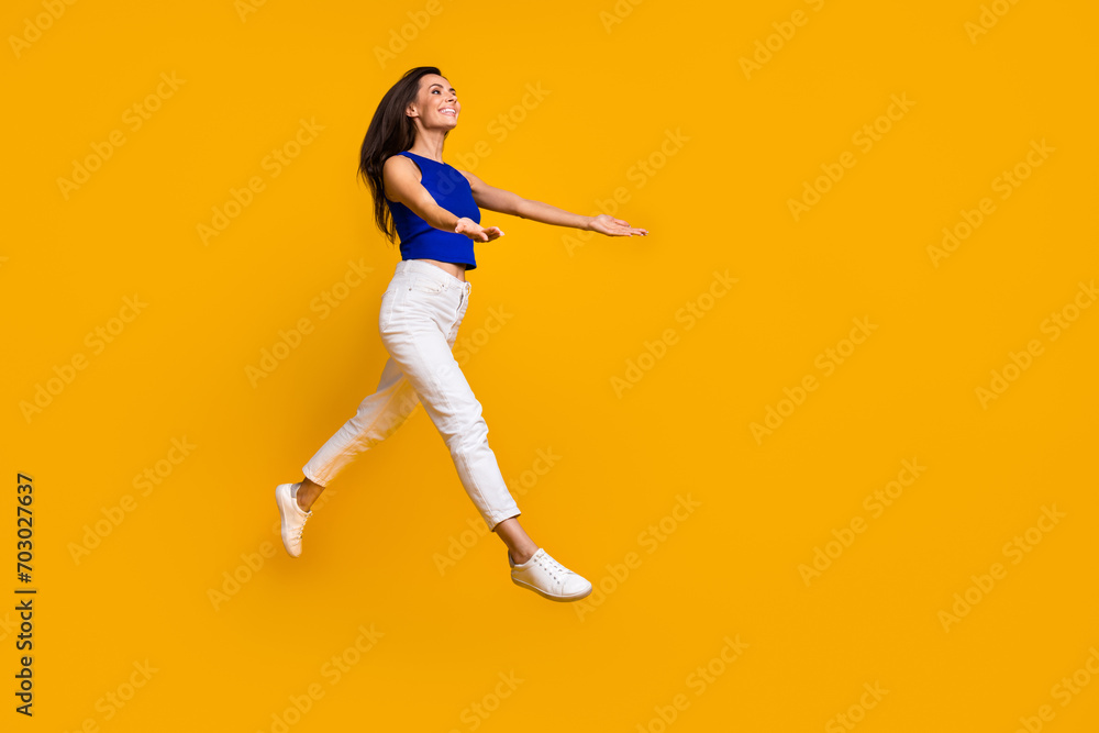Full length photo of pretty excited woman wear blue top jumping holding arms empty space isolated yellow color background