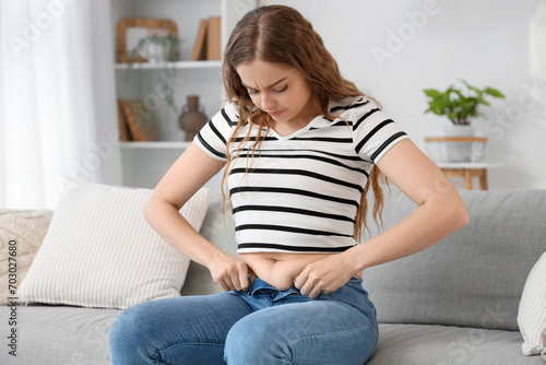 Young woman in tight jeans sitting on sofa at home. Weight gain concept photo