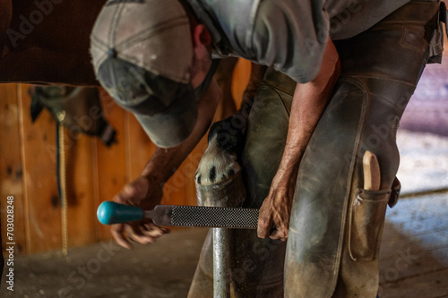 Male farrier trimming bay thoroughbred gelding hoof for shoes in an old rustic barn with wood floors.