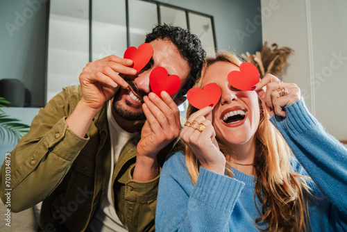 Smiling young couple covering their eyes with red heart shaped papers, having fun at Valentine Day. Boyfriend and girlfriend celebrating their love relationship together on a romantic date at home