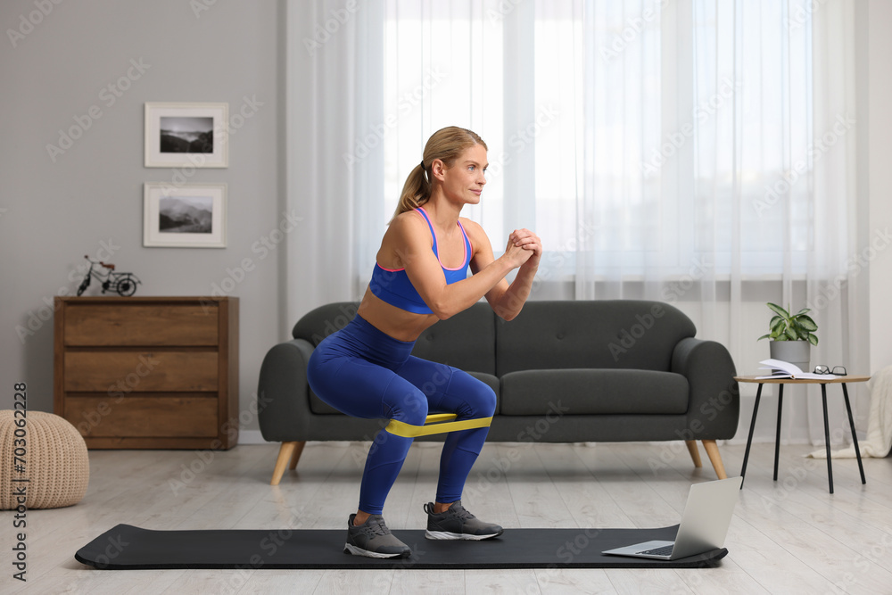 Athletic woman doing squats with fitness elastic band near laptop on mat at home Athletic woman doing squats with fitness elastic band near laptop on mat at home
