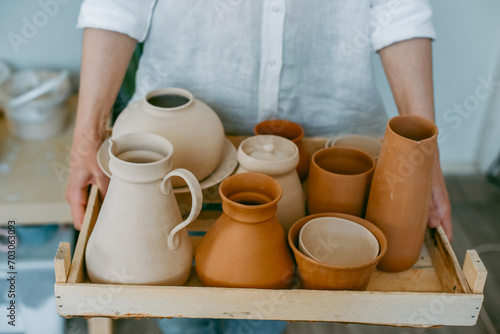 A woman master of a pottery workshop carries clay products for firing. Potter with an assortment of ceramic dishes in a bright workshop © Irina
