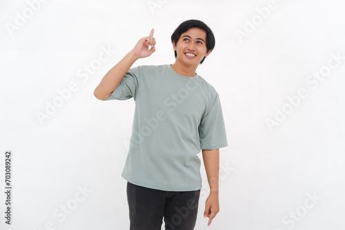Excited Asian male employee wearing a tshirt pointing at the copy space above him, isolated by white background