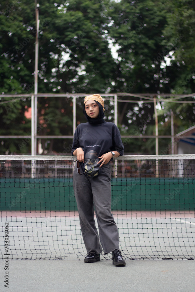 An Asian female model stands confidently on a tennis court, dressed in jeans and a black shirt, adorned with a bandana and glasses