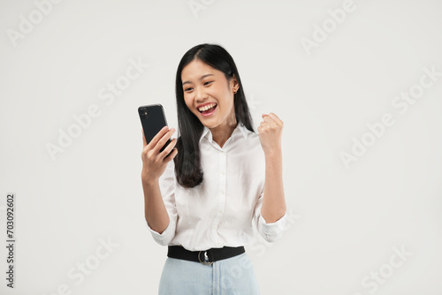 Portrait of a young Asian woman holding her phone feeling excited and celebrating, wearing a white t-shirt and isolated on a white background. 