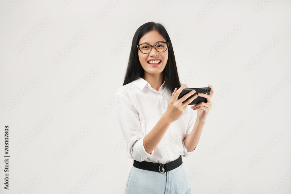 This portrait of a young Asian woman wearing a white shirt and glasses, with a positive expression playing video games on a modern smartphone, isolated on a white background. Modern technology concept This portrait of a young Asian woman wearing a white shirt and glasses, with a positive expression playing video games on a modern smartphone, isolated on a white background. Modern technology concept